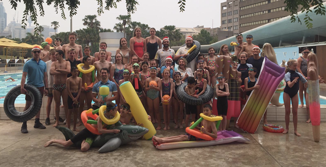 Large group of mixed aged children and adults, standing in front of an outdoor pool with floating water toys.