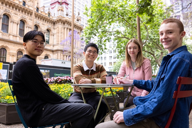Four people sit at an outdoor table in an urban setting with historic buildings and greenery in the background.