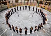 A marching band stands in a circular formation on a patterned pavement, each member holding a brass instrument.