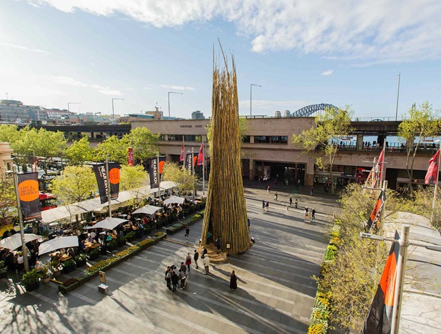 A tall, pointed bamboo sculpture stands in Customs House Square surrounded by people. Nearby are tables, umbrellas, and trees. A bridge is visible in the background under a partly cloudy sky.