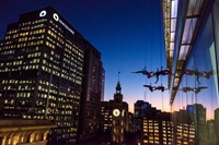 Four people in colourful outfits perform an aerial dance suspended on the side of a glass building at dusk, with city skyscrapers and a clock tower in the background.