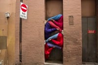 People in colourful clothing are creatively posed within a narrow doorframe next to a "No Entry" sign on a brick wall.
