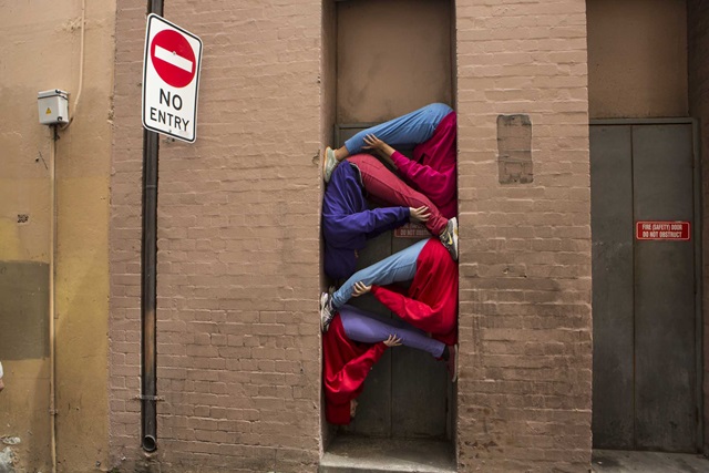 People in colourful clothing are creatively posed within a narrow doorframe next to a "No Entry" sign on a brick wall.
