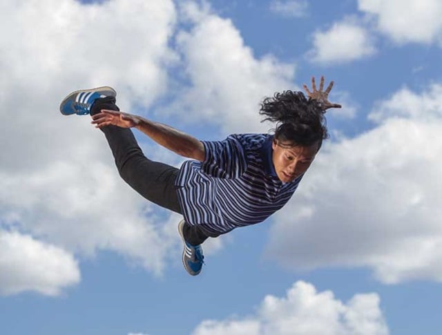 Person in a striped shirt and jeans performs an aerial jump against a background of clouds and blue sky.