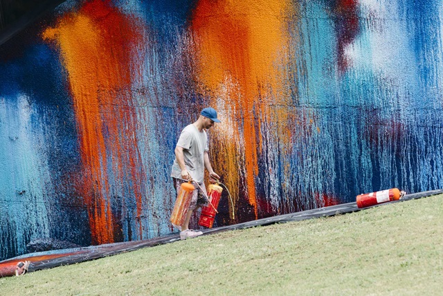 Person carrying paint cans in front of a vibrant, colorful mural on a sloped surface.