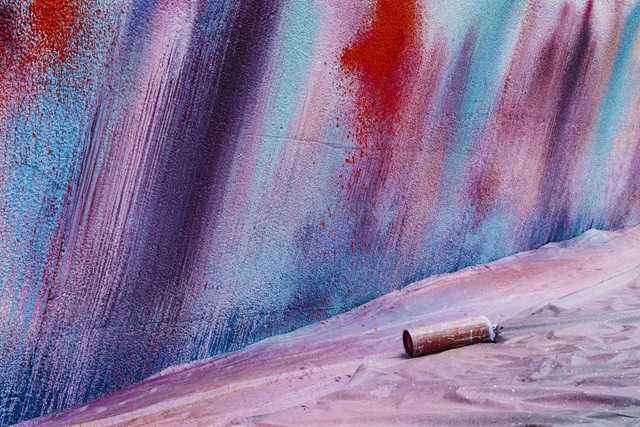A spray-painted wall with vertical streaks in red, blue, and purple hues. A paint can lies on the ground in front.