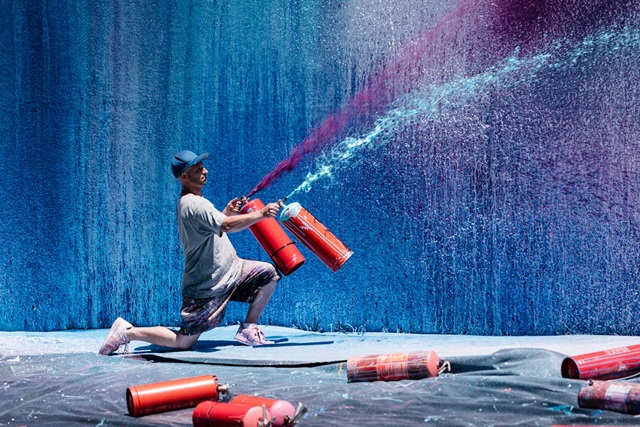 A person sprays blue and purple paint from extinguishers onto a large canvas, with more extinguishers lying on the ground.