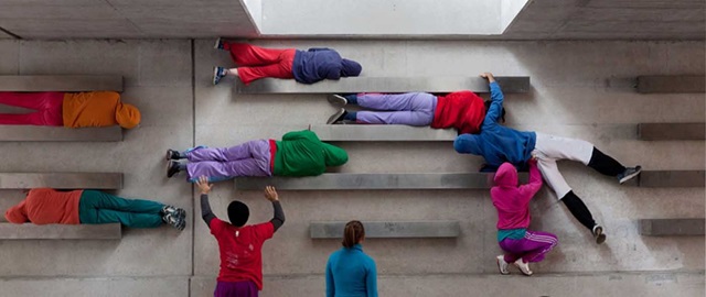 People in colourful clothing lie on elevated beams attached to a concrete wall, with some standing nearby, in an indoor setting.