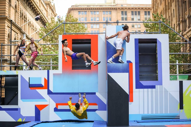 People performing parkour on a colourful urban structure, with one person jumping, others climbing, and another upside down in mid-air. Buildings are visible in the background.