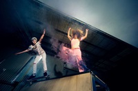 Two skateboarders perform tricks, one on a ramp and the other mid-air, surrounded by colored smoke in an indoor skate park.