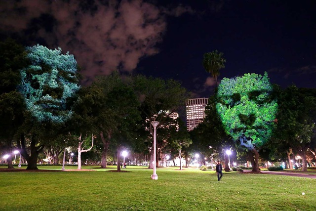 Faces projected onto trees in a park at night, with city buildings in the background and a person walking in the foreground.