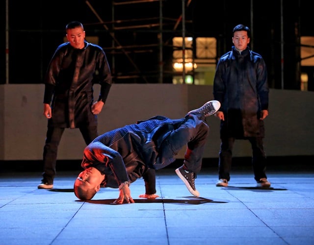 A breakdancer performs a one-handed freeze on a tiled floor, balancing with legs in the air, while two others in dark clothing stand in the background.