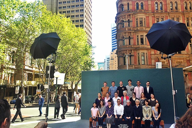 A group of people is posing for a photo in an urban setting with tall buildings, trees, and professional lighting equipment.