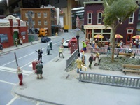 Miniature model city scene with people, buildings, and vehicles; features a red telephone box, a park area with benches, and a corner shop with yellow and red umbrellas.