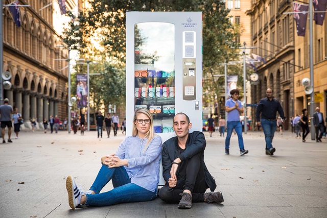 Two people sit on a city street in front of a vending machine surrounded by pedestrians and tall buildings.