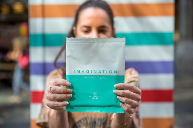 A person holds a teal and white "Imagination" pouch labeled as creative energy fuel, in front of a colorful striped background.
