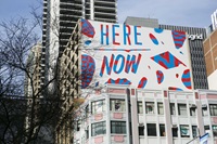 Colourful street art on a building with the words "Here Now" in red and blue. Skyscrapers and trees surround the artwork.