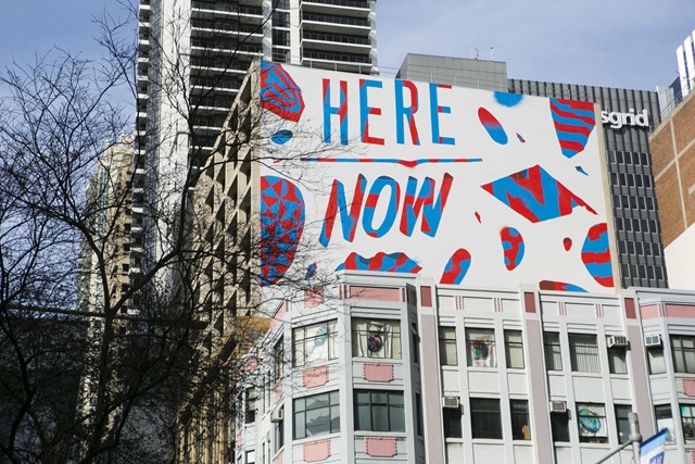 Colourful street art on a building with the words "Here Now" in red and blue. Skyscrapers and trees surround the artwork.