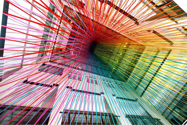Colourful strings are stretched across a large atrium, forming a rainbow pattern converging at the ceiling, surrounded by a modern building structure with windows.