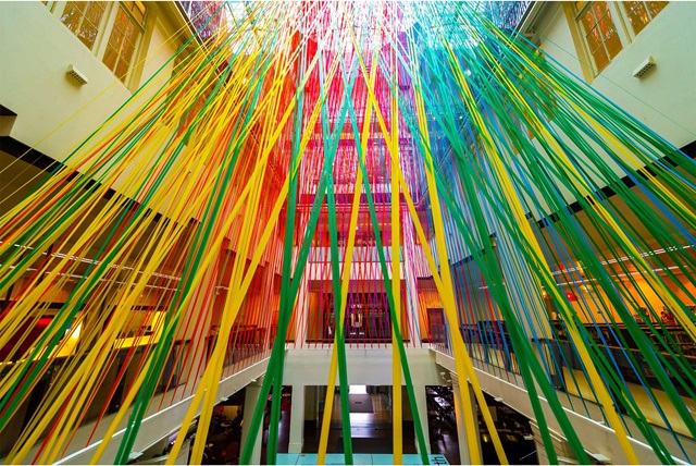 Colourful ribbons hang from the ceiling in an atrium, creating a vibrant canopy effect over the space.