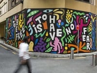 Colourful street mural with abstract shapes and figures. The text reads, "TO BE FREE IS TO HAVE NO FEAR." A person in motion walks past the artwork.