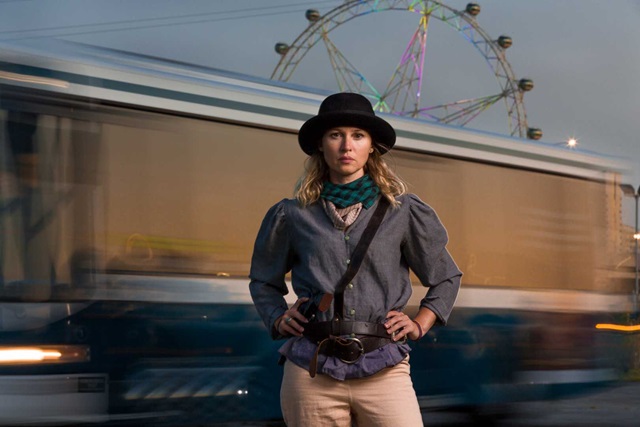 Person wearing a hat and shirt stands with arms at sides in front of a moving bus and a Ferris wheel.