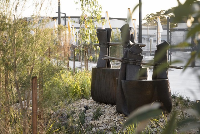 A bronze fountain sculpture resembling two oversized bangala, baskets for carrying water made from palm fronds. They are in a rocky area surrounded by foliage, and there is a paved area with umbrellas in the background.