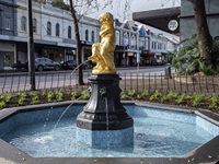 Golden cherub statue on top of cast iron water fountain. Water trickling out of the urn lion themed features on the cast iron fountain.  Street and row houses in the background.