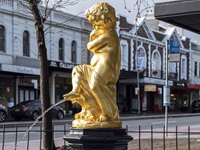 closeup detail of golden cherub statue and water trickling out and urn. Bus sign, street and row houses in the background.
