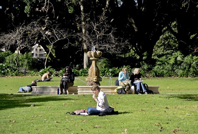 People eat lunch while sitting on the rim and in front of a fountain with a sandstone fish sculpture in its centre. The fountain is on a lawn in a park.