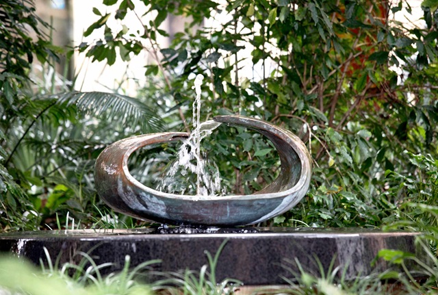A bowel shaped copper fountain spurting water on a stone base set amongst parkland