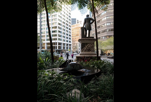 A fountain set among trees and greenery with large statue background, set against a city backdrop