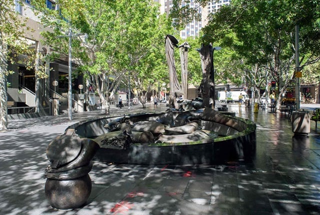 Large ornate fountain in a pedestrian area surrounded by trees and street furniture