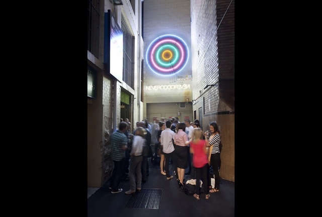 A crowd gathers in an atrium. On the wall above them is a neon sign of concentric circles, below which is another neon sign reading "NEVERODDOREVEN".
