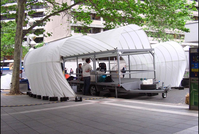 large white tent style shelter fixed to a trailer houses a D.J. and other equipment amongst city buildings