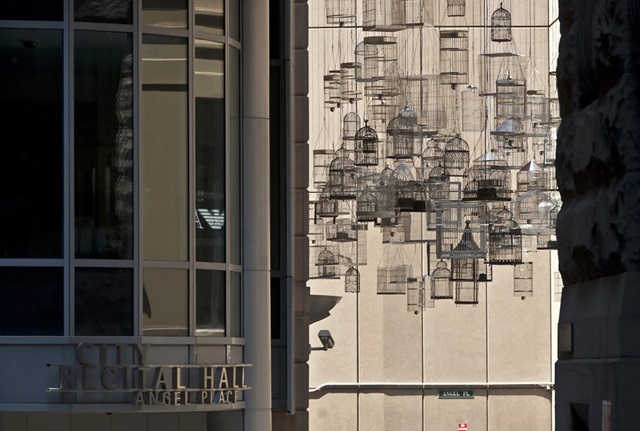 A large number of empty birdcages hang in a laneway next to City Recital Hall, above a street sign reading Angel Place.