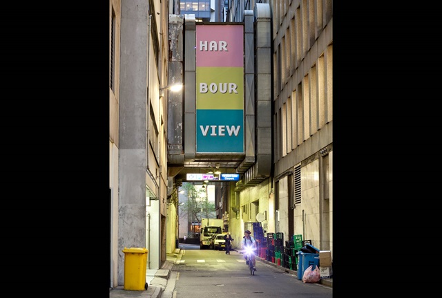 A large text banner fixed to the wall of the elevated brick corridor joining the buildings on either side of narrow lane.