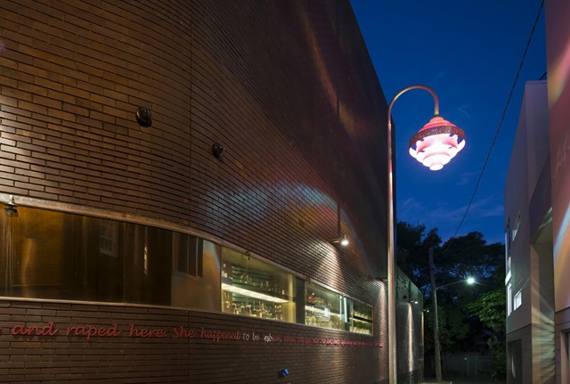 A streetlight with a pink decorative lampshade is beside a curved brick building. Pink, handwriting-style text on the side of the building reads, "...and raped here. She happened to be lesbian..."