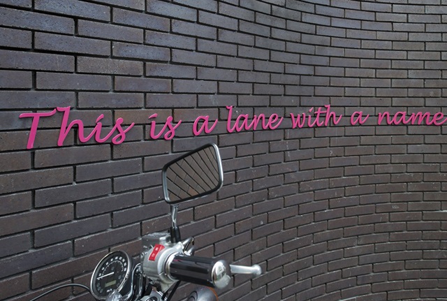 A brick wall with pink, handwriting-style text on it reading, "This is a lane with a name..." The handlebar of a motorbike is in front of the wall, below the text.