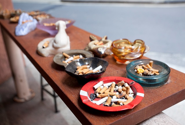 Outdoor wooden bar counter with black bar stool standing beside it with 12 ashtrays in a variety of shapes sitting on top.