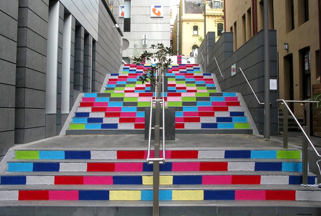 An outdoor staircase in the city is painted along the front of the steps with brightly coloured rectangles, creating a mosaic effect up the stairs.