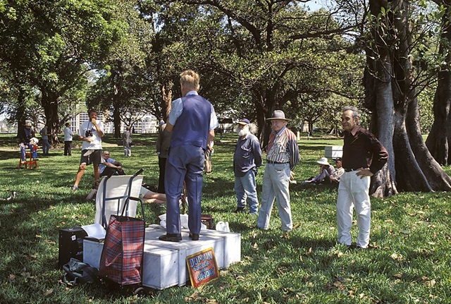 A man stands on a marble soapbox in The Domain on a sunny day, while about a dozen other people look on. There are several bags and signs around the soapbox, one of which reads, "Domain soapbox every Sunday".
