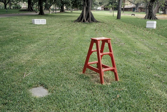 A bright red stepladder stands on a grassy lawn near a plaque. In the distance are two marble sculpture resembling old-fashioned soapboxes, several trees, and a footpath.