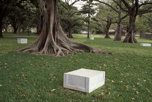 A marble sculpture resembling a wooden crate rests on a grassy lawn. In the distance are three similar marble sculptures and several trees.