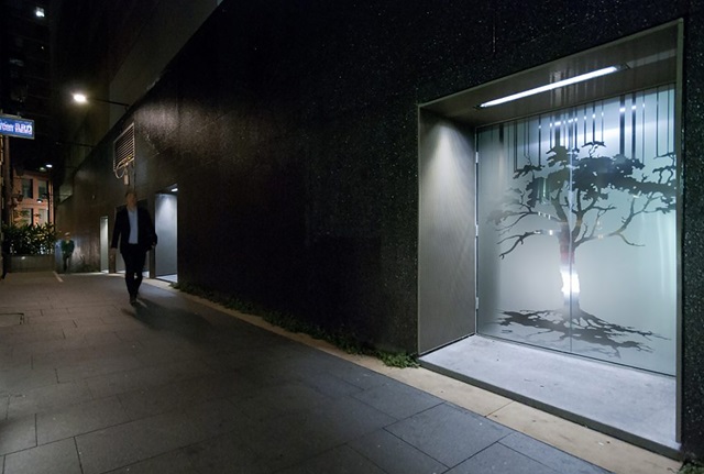 Albion Place at night. An illuminated doorway displays a steel double door etched with a silhouette of a tree. A pedestrian walks down the laneway towards the doorway.