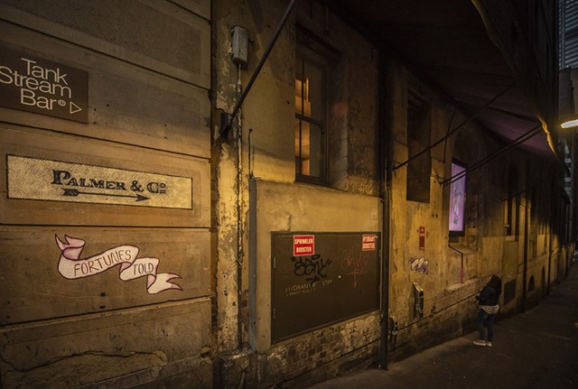 A laneway at evening. On the sandstone wall of a building is a painted ribbon, on which are the handwritten words, "Fortunes told". Above it is an old-fashioned painted sign reading "Palmer & Co." is an arrow pointing down the laneway, and above that is a modern sign reading "Tank Stream Bar". Further down the laneway, a pedestrian looks up at a video screen with the image of a ghost.