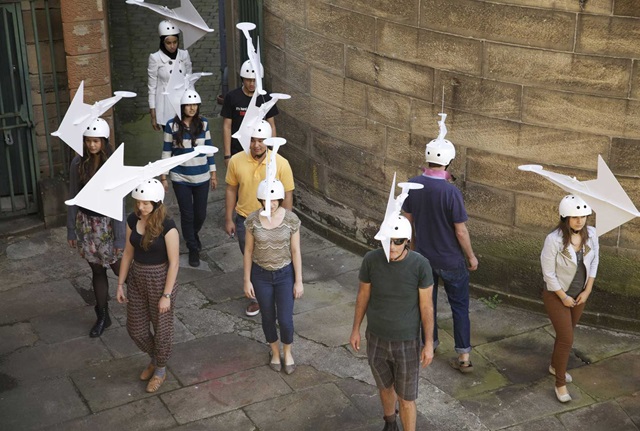 10 people stand in a stone walled area wearing white helmets with wind weathervanes  attached to the top.