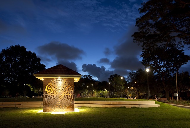 A small, square brick building on a lawn in a park in late evening. The building is decorated with spiderweb-like aluminium grills, and illuminated.