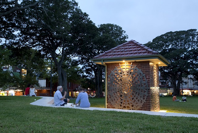 A small, square brick building on a lawn in a park in early evening. The building is decorated with spiderweb-like aluminium grills, and illuminated. Two people sit on concrete stairs nearby, having a conversation and sharing food.