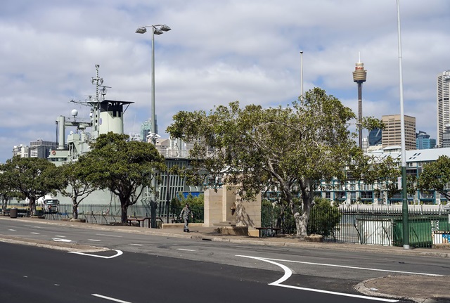 A soldier in camouflage walks on the footpath past a sandstone memorial. The street is lined with trees and runs along the shore. In the background is a large naval ship.